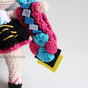 May include: Crocheted doll with pink, blue, and black clothing. The doll has a yellow and black striped skirt and a blue bow. The doll is holding a pink and blue crocheted object.