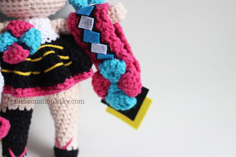 May include: Crocheted doll with pink, blue, and black clothing. The doll has a yellow and black striped skirt and a blue bow. The doll is holding a pink and blue crocheted object.