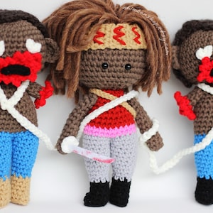 May include: Three crocheted dolls with brown skin, red lips, and white eyes. The doll in the middle has brown hair and is wearing a red, pink, and gray outfit. The dolls on either side are wearing blue pants and brown shoes.