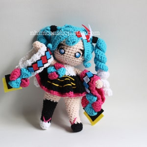 May include: Crocheted anime character doll with blue hair, a white and red headband, a black and yellow skirt, and pink and blue accessories. The doll is standing on a white background.