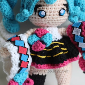 May include: Crocheted amigurumi doll with blue hair, a pink and blue bow, a black and yellow striped skirt, and a white, pink, and blue crocheted jacket. The doll is wearing a white crocheted dress underneath the jacket. The doll is standing on a white surface. The text "nhinconshop.etsy.com" is printed on the doll's skirt.