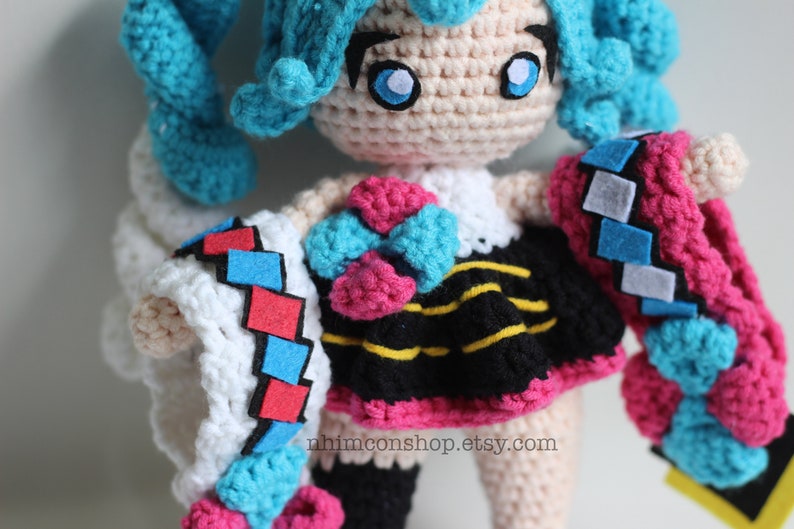 May include: Crocheted amigurumi doll with blue hair, a pink and blue bow, a black and yellow striped skirt, and a white, pink, and blue crocheted jacket. The doll is wearing a white crocheted dress underneath the jacket. The doll is standing on a white surface. The text "nhinconshop.etsy.com" is printed on the doll's skirt.