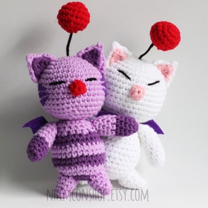 May include: Two crocheted plush toys, one purple and one white, with red pom-poms on top of their heads. Both toys have cat-like features and are standing side-by-side.