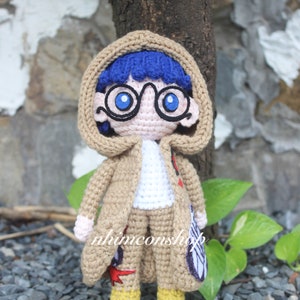 Puede incluir: Muñeca amigurumi de crochet con un abrigo marrón, una camisa azul y gafas azules. La muñeca tiene el pelo azul y está de pie sobre una superficie gris.