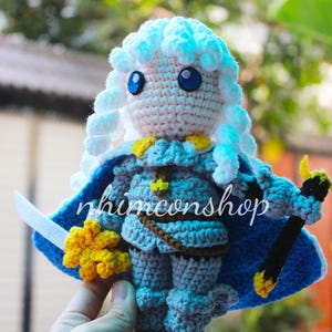 Könnte beinhalten: Gehäkelte Puppe einer Figur mit blauen Augen, weißen Haaren und einem blauen Umhang. Die Puppe trägt eine graue Rüstung mit gelben Akzenten und hält ein Schwert und eine gelbe Blume. Der Text "nhimconshop" ist auf dem Umhang zu sehen.