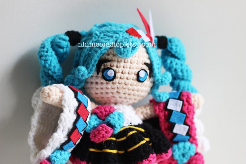 May include: Crocheted plush toy of a female anime character with blue hair in pigtails, wearing a white, black, pink, and blue outfit. The toy has large blue eyes and a white face. The text "nhimconshop.etsy.com" is visible on the toy's chest.