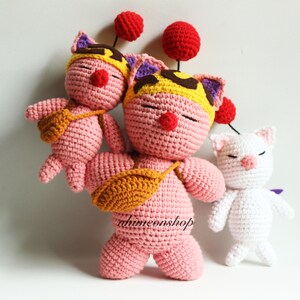 May include: Three crocheted plushies: a pink one with a yellow and purple hat, a smaller pink one with a yellow hat, and a white one with a pink nose. All three have red pom-poms on their heads.