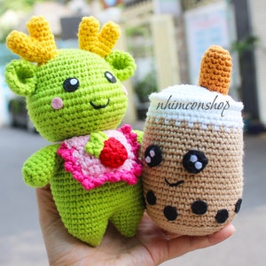 May include: Two crocheted plush toys. One is a green reindeer with yellow antlers and a pink bib. The other is a brown crocheted boba tea with a white top and black boba pearls. The text "nhimconshop" is on the boba tea.