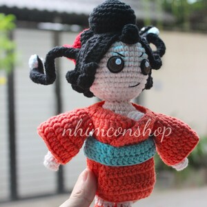 Puede incluir: Mu&ntilde;eca de ganchillo hecha a mano con cabello negro peinado en mo&ntilde;o y rizos laterales. La mu&ntilde;eca lleva un kimono rojo con una faja turquesa. La cara de la mu&ntilde;eca est&aacute; detallada con ojos azules y una expresi&oacute;n sonriente. El texto "nhimconshop" est&aacute; en el kimono.