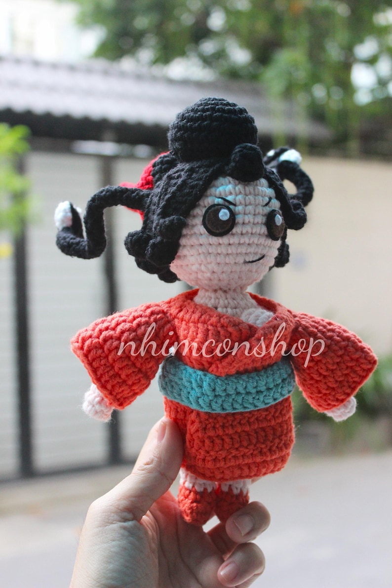 Puede incluir: Mu&ntilde;eca de ganchillo hecha a mano con cabello negro peinado en mo&ntilde;o y rizos laterales. La mu&ntilde;eca lleva un kimono rojo con una faja turquesa. La cara de la mu&ntilde;eca est&aacute; detallada con ojos azules y una expresi&oacute;n sonriente. El texto "nhimconshop" est&aacute; en el kimono.