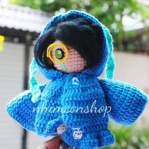 May include: A handmade crochet doll with a blue hooded jacket and black hair. The doll has a single eye with a yellow frame and a green iris. The text "nhimconshop" is visible on the jacket.