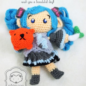 May include: A crocheted doll with blue hair, a grey and blue dress, and orange bear. The doll is holding the bear in its left hand and a white carrot in its right hand. The doll is wearing black shoes. The doll is sitting on a white surface. The text "wish you a beautiful day!" is written above the doll.