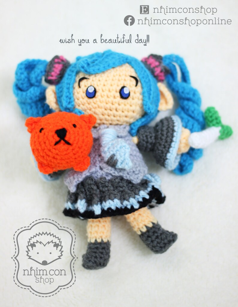 May include: A crocheted doll with blue hair, a grey and blue dress, and orange bear. The doll is holding the bear in its left hand and a white carrot in its right hand. The doll is wearing black shoes. The doll is sitting on a white surface. The text "wish you a beautiful day!" is written above the doll.