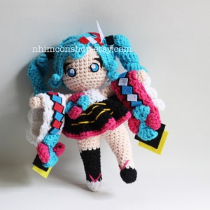May include: Crocheted plush toy of a blue-haired anime character with a pink and black dress, white and blue sleeves, and yellow accents. The toy is holding a blue and white crocheted scarf.