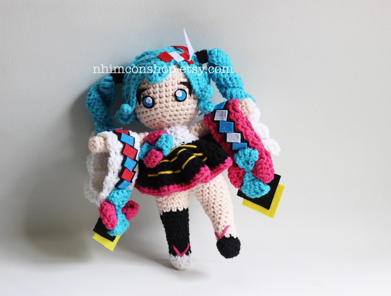 May include: Crocheted plush toy of a blue-haired anime character with a pink and black dress, white and blue sleeves, and yellow accents. The toy is holding a blue and white crocheted scarf.