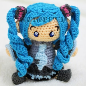 May include: A crocheted blue-haired doll with pink hair ties, wearing a blue and gray outfit. The doll has large blue eyes and a light-colored face. The doll is sitting on a white surface. The text "NHIMCONSHOP.ETSY.COM" is visible on the doll's chest.