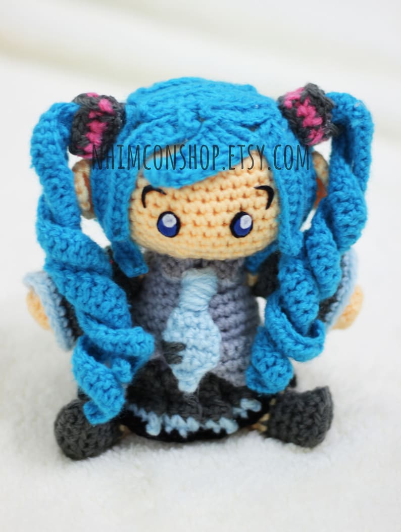 May include: A crocheted blue-haired doll with pink hair ties, wearing a blue and gray outfit. The doll has large blue eyes and a light-colored face. The doll is sitting on a white surface. The text "NHIMCONSHOP.ETSY.COM" is visible on the doll's chest.