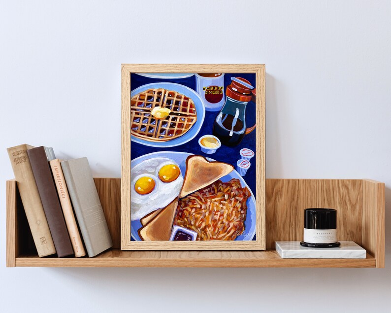 May include: A framed painting of a breakfast plate with eggs, toast, and hash browns, alongside a waffle with butter, syrup, and coffee. The painting is set against a blue background, with a wooden frame.