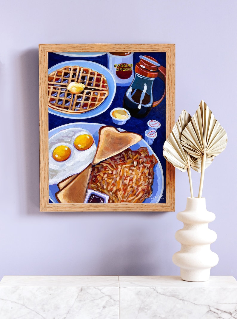 May include: A framed painting of a breakfast scene. The painting depicts a waffle with butter, eggs, toast, and hash browns on a blue plate. A bottle of syrup and small containers of condiments are also visible. The painting is in a wooden frame.