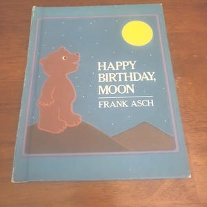 May include: A children's book titled "Happy Birthday, Moon" by Frank Asch. The cover features a brown bear silhouette, a yellow moon, and a starry night sky. The book has a blue border.