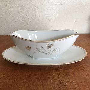 May include: White porcelain gravy boat with a gold trim and a delicate floral design. The boat is on a matching saucer.