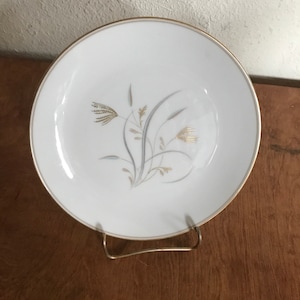 May include: A white ceramic plate with a gold rim and a gold and gray floral design. The plate is displayed on a gold-colored metal stand. The design features stylized wheat or grass.