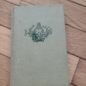 May include: A vintage book with a pale green cover. The cover has a green illustration of two figures near a structure, framed by leafy branches. The book is on a wooden surface.