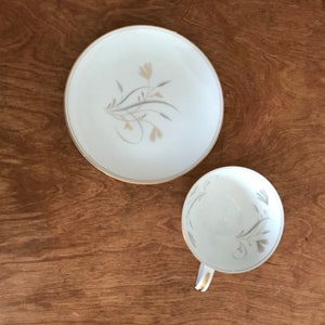 May include: A white teacup and saucer set with a gold rim and a floral design in gray and beige. The teacup has a handle and is placed next to the saucer on a wooden surface.