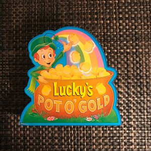 May include: A cartoon leprechaun holding a pot of gold with a rainbow above him. The pot is labeled "Lucky's Pot O' Gold".