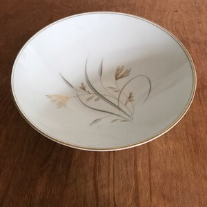 May include: A white ceramic plate with a gold rim and a floral design in shades of gray and gold. The plate has a shallow bowl shape and is set against a wooden surface.