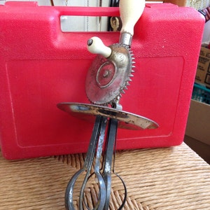 May include: A vintage hand-crank egg beater with a white handle and a metal whisk attachment. The beater is resting on a red plastic container.