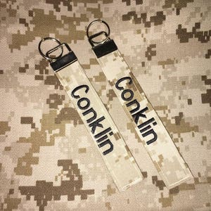May include: Two tan and brown camouflage keychains with black embroidered text that reads "Conklin".