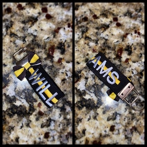 May include: Two black and yellow striped keychains with silver metal rings. One keychain has the word "WILL" embroidered in silver thread. The other keychain has the word "AMS" embroidered in silver thread.