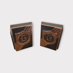 May include: Two bronze-colored cufflinks with a shield design and the letter "D" in the center. The cufflinks have a rectangular shape with a dark background and bronze accents. The design is symmetrical.
