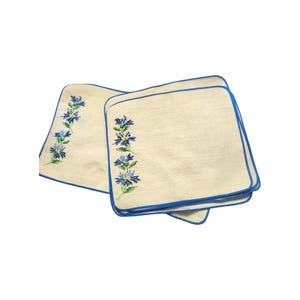 May include: A set of square off-white fabric napkins featuring a blue border and a vertical cross-stitch pattern of blue cornflowers with green stems and leaves. The napkins are stacked, displaying the floral embroidery on the left side.