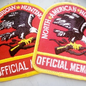 May include: Two embroidered patches with a red background and yellow border. Each patch features a black and white eagle clutching a branch, with the text "NORTH AMERICAN HUNTING CLUB" and "OFFICIAL MEMBER" in white.