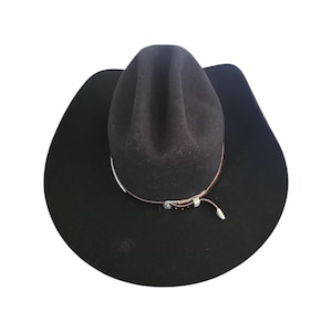 May include: A black felt cowboy hat featuring a tall, creased crown and a wide, curved brim. A thin, dark brown leather hat band wraps around the base of the crown, secured with a small silver-toned metal buckle and a dangling leather strap end.