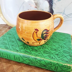 May include: A ceramic mug with a rooster design. The mug is tan with a brown interior and a curved handle. The rooster is yellow, red, and green. The mug is sitting on a green book.