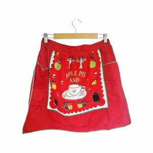 May include: Red apron with a white and green decorative border. The apron features a central design with the words "APPLE PIE AND" above a teacup and saucer, surrounded by apples and trees. A black teapot is also visible.