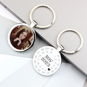 May include: Silver keyring with a round photo frame and a calendar disc. The photo frame shows a woman with brown hair. The calendar disc shows the month of November 2023.
