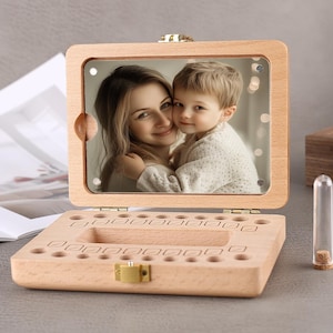 May include: A wooden baby tooth keepsake box with a hinged lid and a clear acrylic window displaying a photo of a mother and child. The box has individual slots for each tooth and a small glass vial.