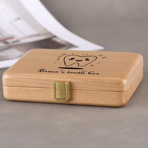 May include: A light-colored wooden tooth box with a gold-colored clasp. The box is rectangular and has a smiling tooth graphic and the text "Bruce's tooth box" engraved on the lid.
