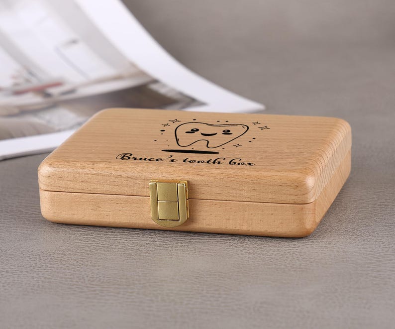 May include: A light-colored wooden tooth box with a gold-colored clasp. The box is rectangular and has a smiling tooth graphic and the text "Bruce's tooth box" engraved on the lid.