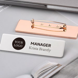 Op de afbeelding: Een wit rechthoekig naambordje met een zwarte cirkel waarop "YOUR LOGO" staat en de tekst "MANAGER Krista Brantly". Het naambordje is bevestigd aan een rozégouden metalen pin.