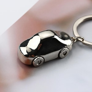 K&ouml;nnte beinhalten: Ein gl&auml;nzender, silberner Autoschl&uuml;sselanh&auml;nger. Das Miniaturfahrzeug hat vier R&auml;der und ist an einem silbernen Schl&uuml;sselring befestigt. Die Oberfl&auml;che des Autos reflektiert das Licht und erzeugt ein helles, metallisches Aussehen. Ein schlichtes, aber stilvolles Accessoire.