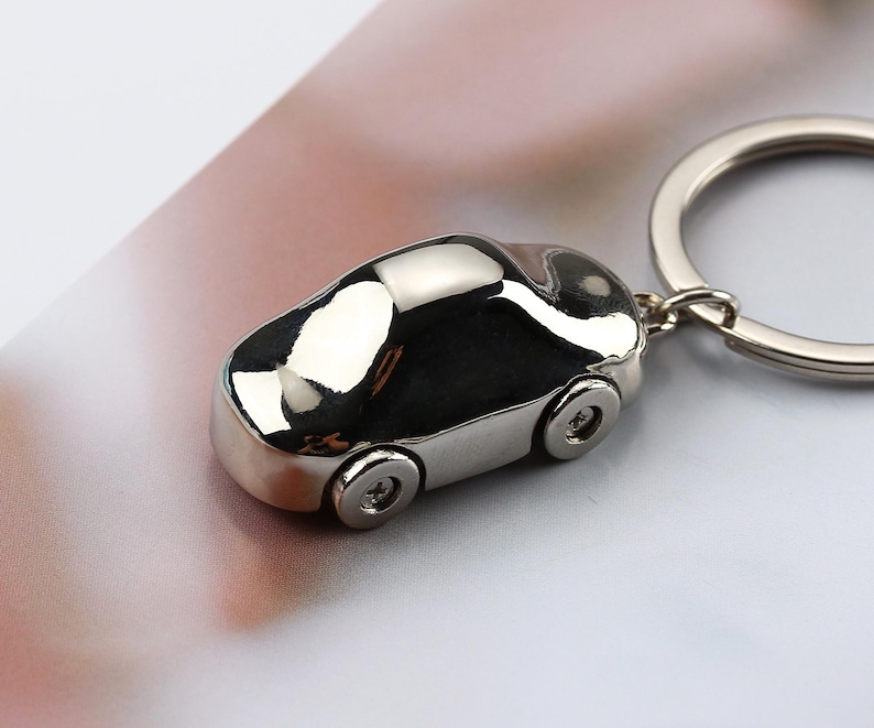 K&ouml;nnte beinhalten: Ein gl&auml;nzender, silberner Autoschl&uuml;sselanh&auml;nger. Das Miniaturfahrzeug hat vier R&auml;der und ist an einem silbernen Schl&uuml;sselring befestigt. Die Oberfl&auml;che des Autos reflektiert das Licht und erzeugt ein helles, metallisches Aussehen. Ein schlichtes, aber stilvolles Accessoire.