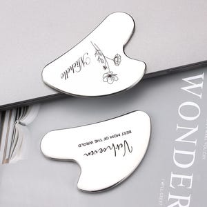 Op de afbeelding: Twee zilverkleurige metalen gua sha-gereedschappen met gegraveerde tekst. Het bovenste gereedschap heeft het woord "Michelle" gegraveerd met een bloemmotief. Het onderste gereedschap heeft de tekst "Verhoeven" en "Best Mom of the World" gegraveerd.