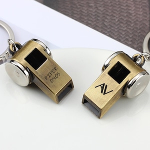 May include: Two bronze-colored whistle keychains with silver accents. One keychain has the text "PIPER 04.05" engraved on it. The other keychain has the initials "AV" engraved on it. Both keychains are on silver keyrings.
