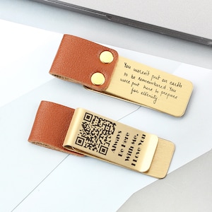Könnte beinhalten: Zwei goldene Lesezeichen-Clips mit Lederriemen. Ein Clip hat ein Zitat über die Ewigkeit und der andere ein Zitat über die Liebe und einen QR-Code.