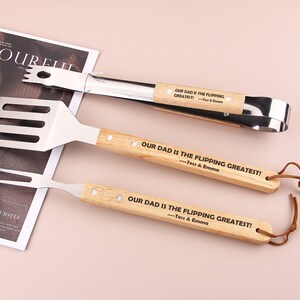 Puede incluir: Un juego de tres herramientas para asar a la parrilla con mangos de madera. Los mangos están grabados con el texto "Nuestro papá es el mejor!" y los nombres "Tess & Emma".